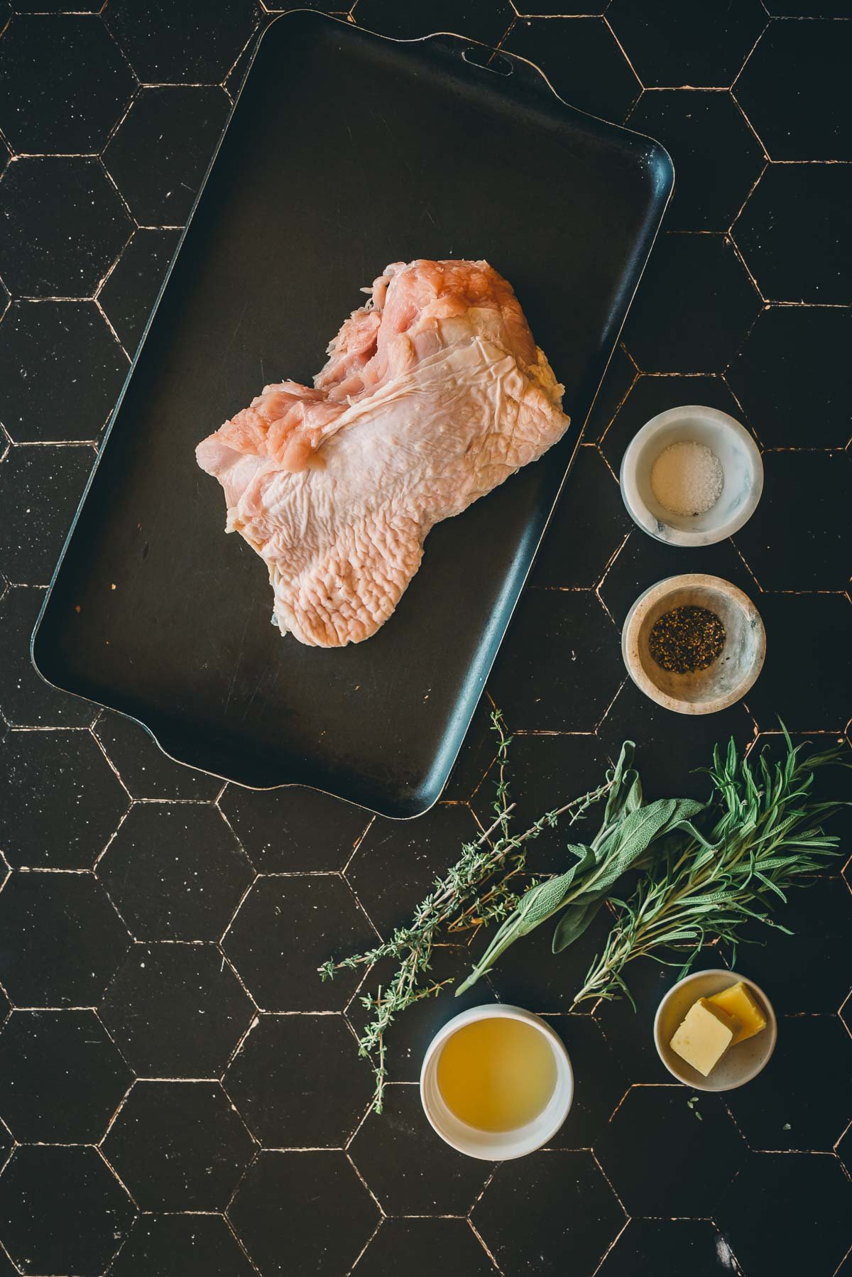 Raw turkey breast on a baking tray with bowls of salt, pepper, butter, olive oil, and sprigs of herbs on a black tiled surface.