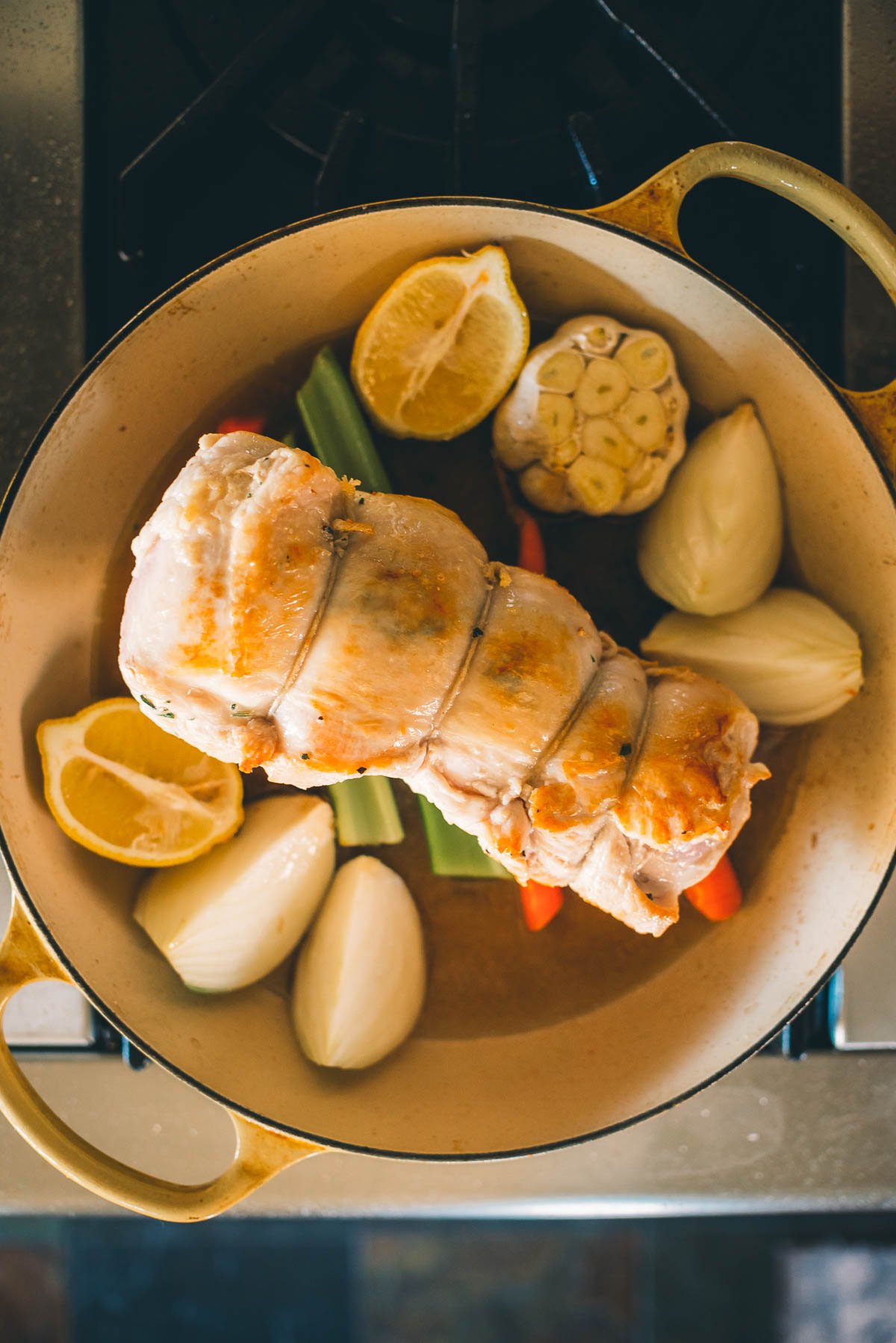 A tied turkey breast roast is in a pot with lemon halves, onions, garlic bulb, and vegetables, ready for cooking.