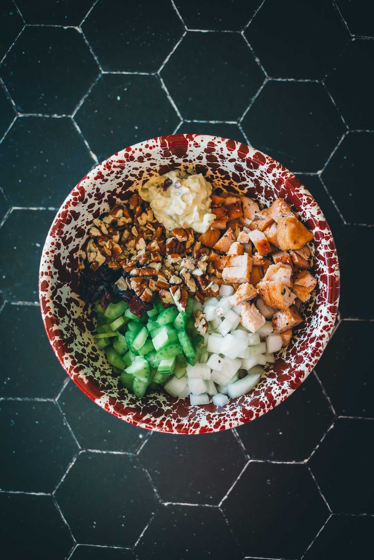 A bowl with leftover turkey meat, chopped celery, onions, nuts, cranberries, and a dollop of mustard, set on a dark tiled surface.