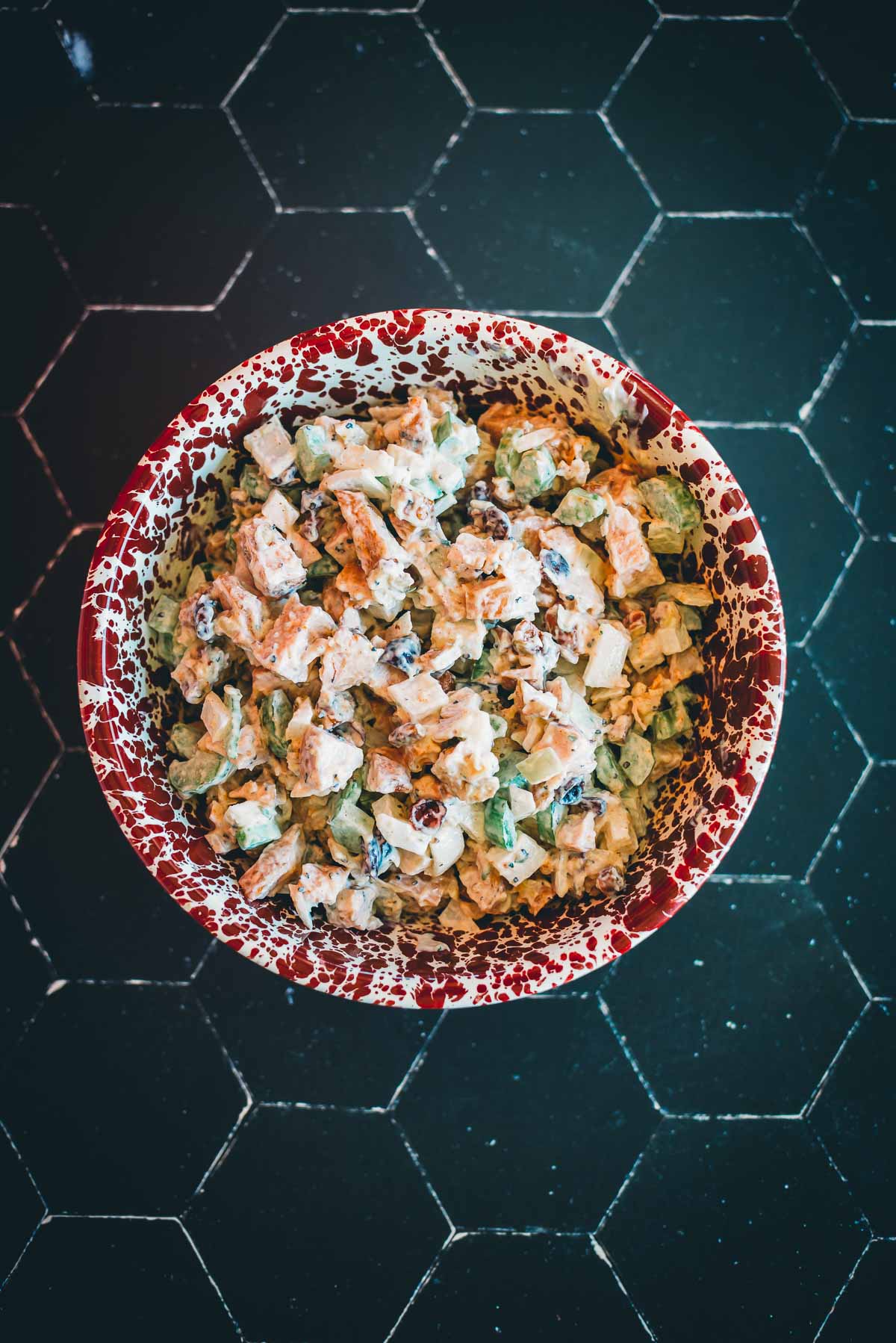 A speckled red and white bowl filled with a leftover turkey salad on a dark tile surface.