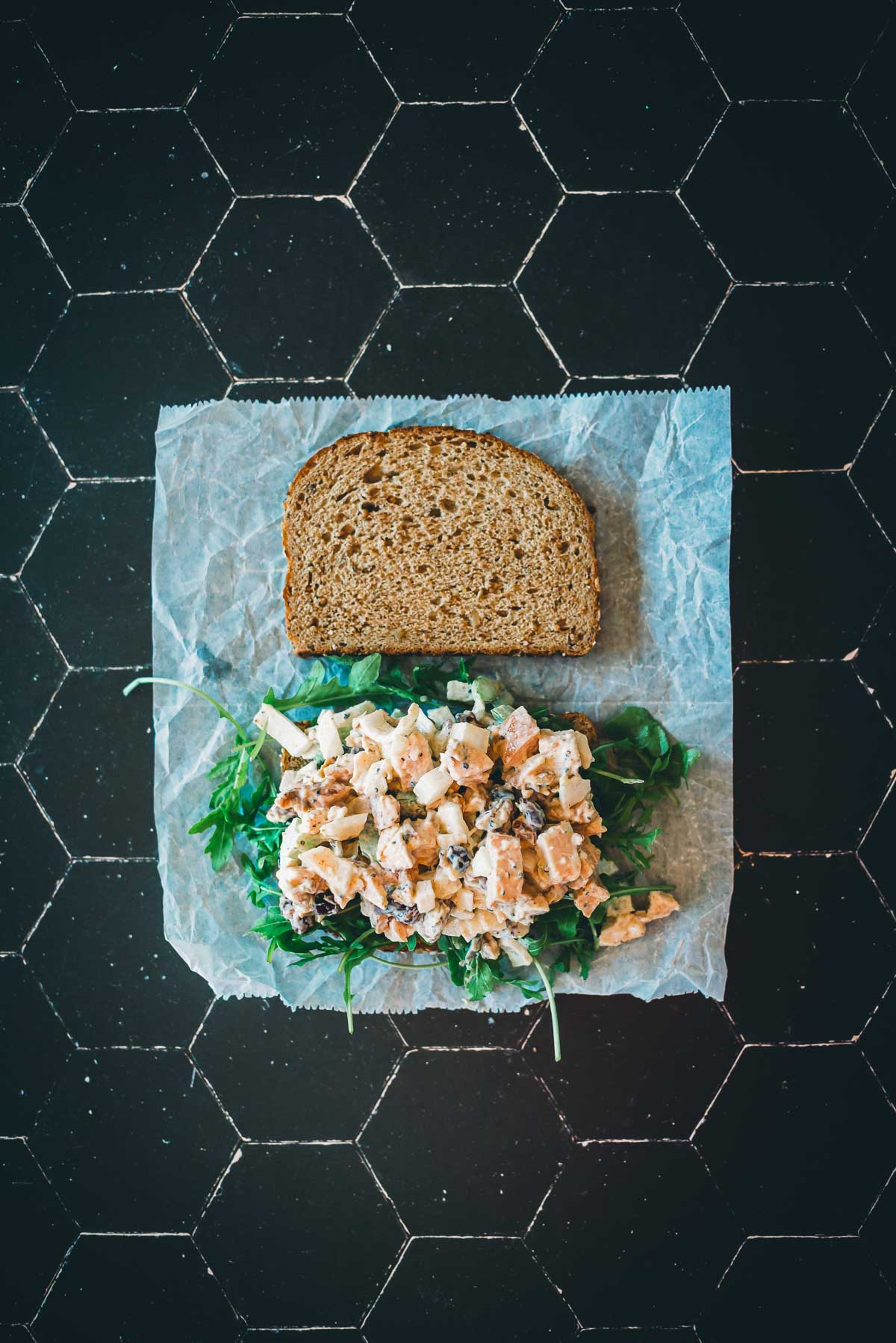 Open-faced sandwich with turkey salad, greens, and a slice of whole grain bread on parchment paper over a black hexagonal tile surface.