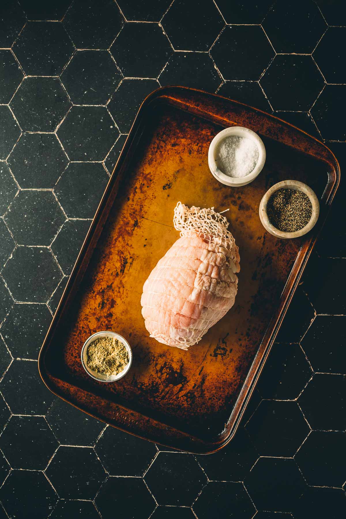 A raw, net-wrapped boneless turkey breast on a baking tray, surrounded by three small bowls containing salt, pepper, and herbs on a dark tiled surface.