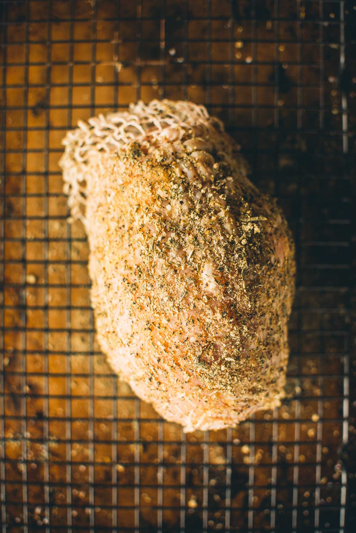 Seasoned boneless turkey breast on a wire rack, covered in herbs and spices, with a netting on one end.