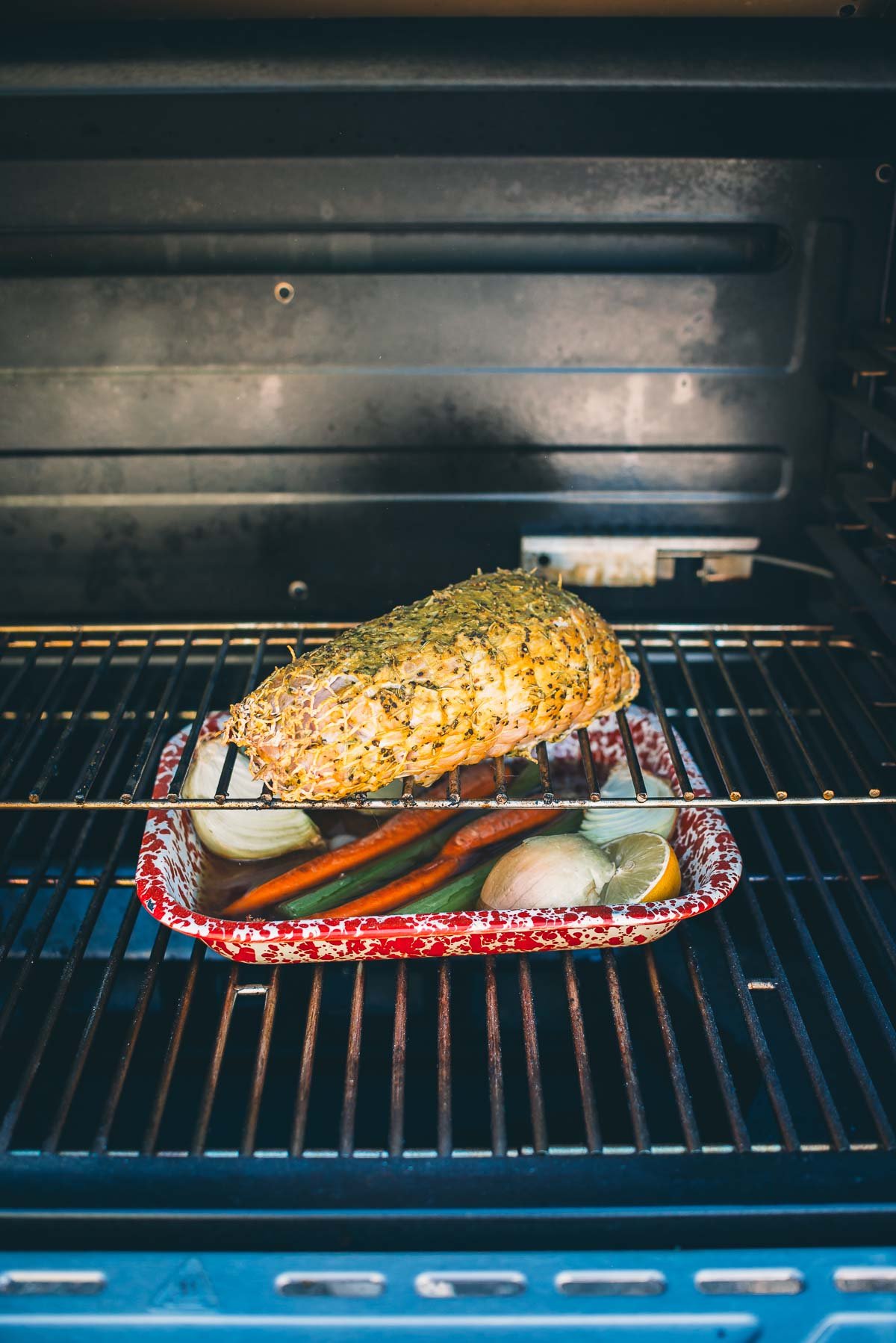 A seasoned boneless turkey breast in a smoker, with onions and carrots in a red roasting pan below.