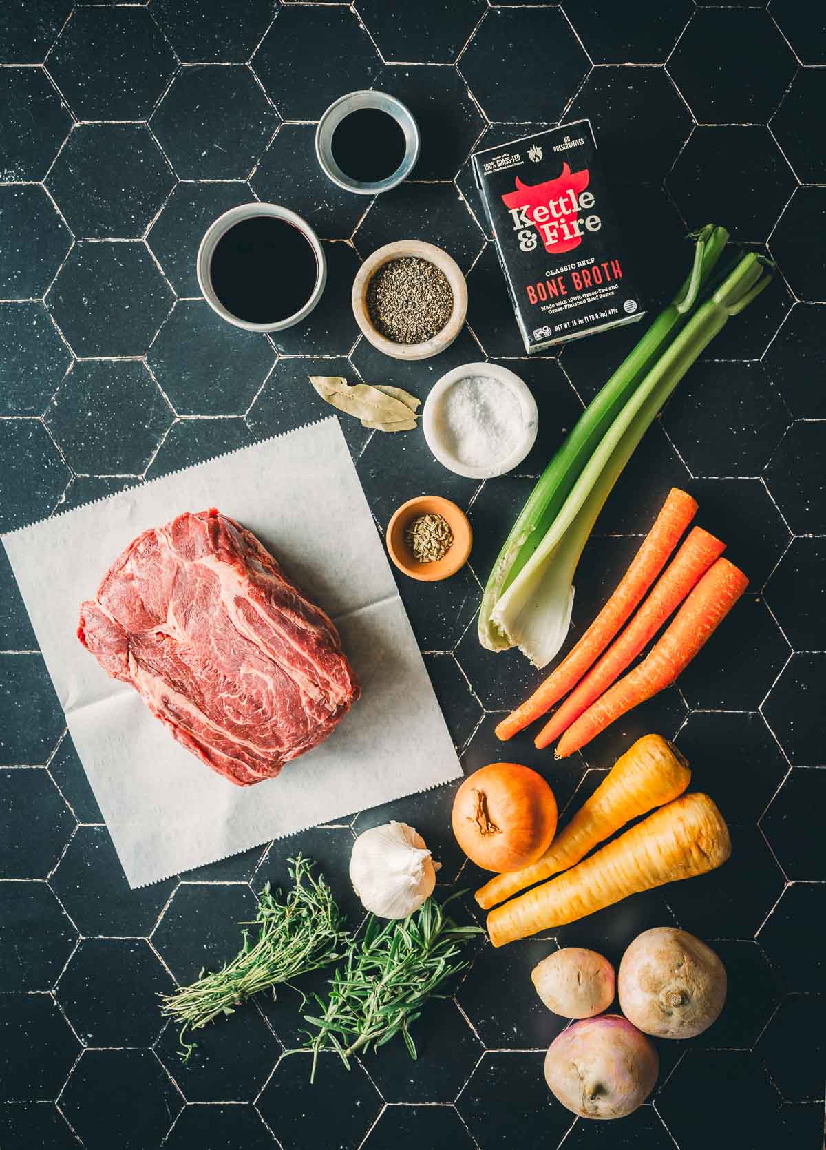 A selection of ingredients on a black tile surface: chuck roast, carrots, celery, potatoes, onion, garlic, rosemary, seasonings, bone broth, and red wine.