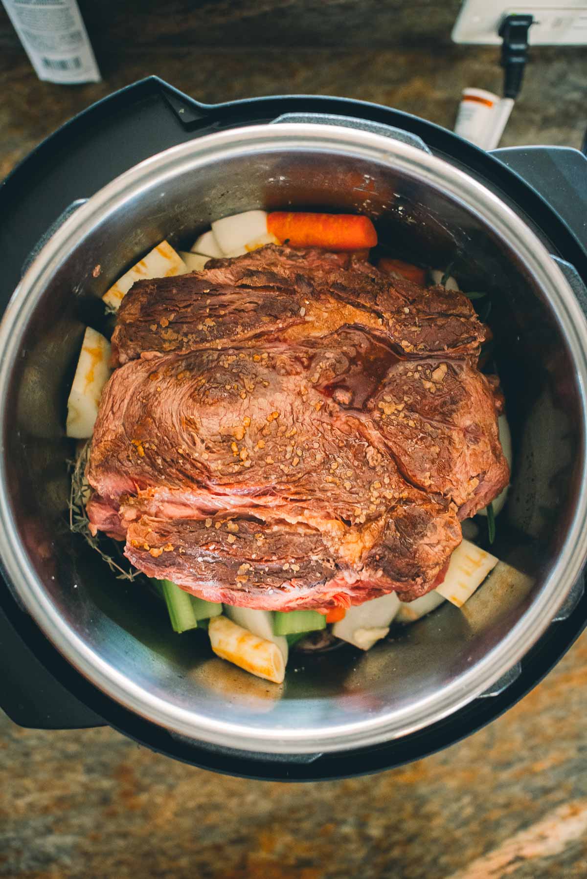 A raw, seasoned chuck roast sits in an Instant Pot with chopped carrots, celery, and parsnips.