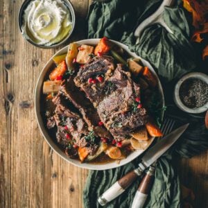 A bowl of instapot chuck roast and vegetables on a wooden table, accompanied by a small dish of whipped cream and a knife and fork.