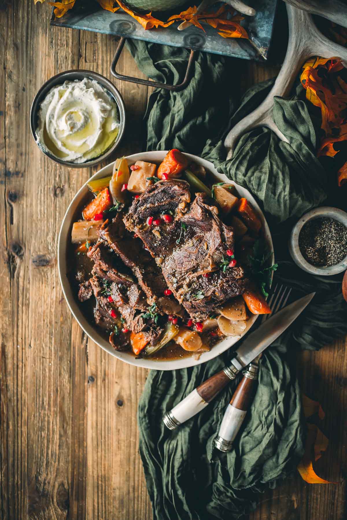 A bowl of instapot chuck roast and vegetables on a wooden table, accompanied by a small dish of whipped cream and a knife and fork.