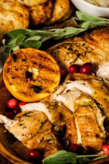 Sliced roast turkey with herbs, grilled orange slices, and cranberries on a wooden platter. Gravy in a white dish and roasted potatoes in the background.