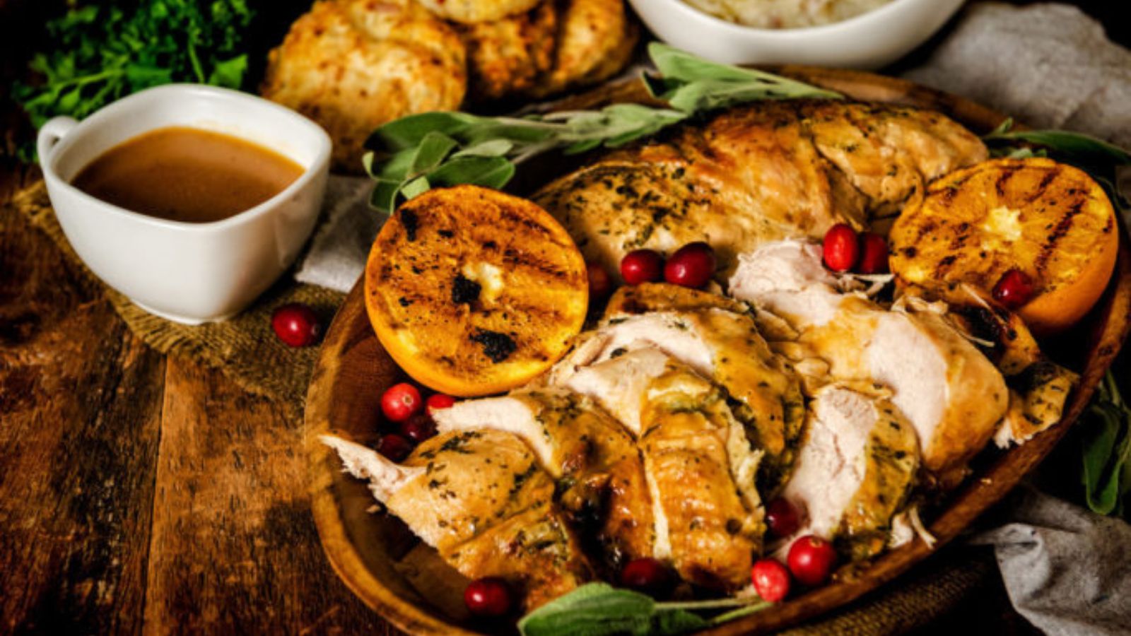 Sliced roast turkey with herbs, grilled orange slices, and cranberries on a wooden platter. Gravy in a white dish and roasted potatoes in the background.