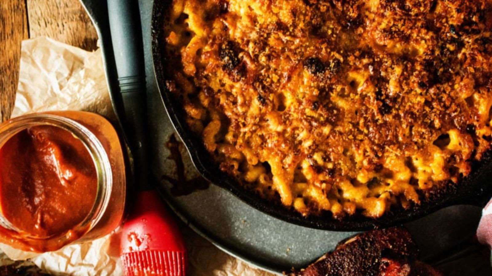 Skillet of baked macaroni and cheese with a crispy topping, alongside a jar of barbecue sauce and a red basting brush on a wooden table.