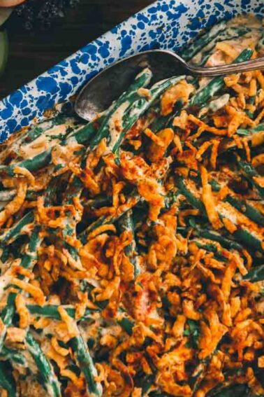 A blue speckled dish filled with green bean casserole topped with crispy fried onions, served with a silver serving spoon.