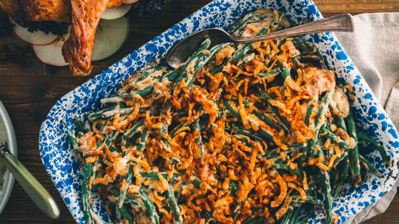 A blue speckled dish filled with green bean casserole topped with crispy fried onions, served with a silver serving spoon.