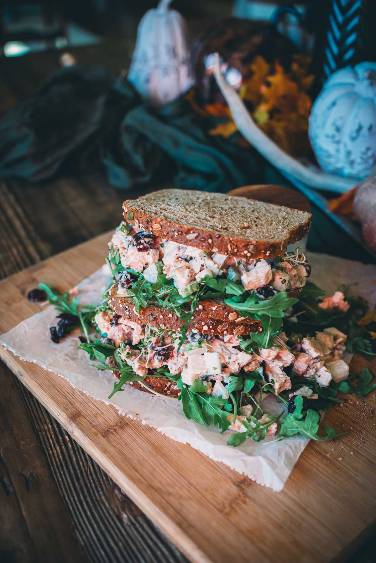 A stacked turkey salad sandwich with wheat bread, greens, and a salad mix on a wooden board with fall-themed decorations in the background.