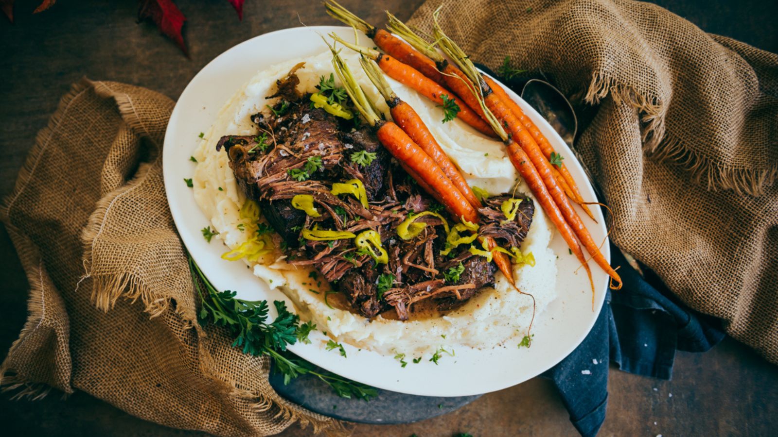 A plate with shredded beef on mashed potatoes, garnished with carrots, herbs, and green peppers, on a rustic burlap background.