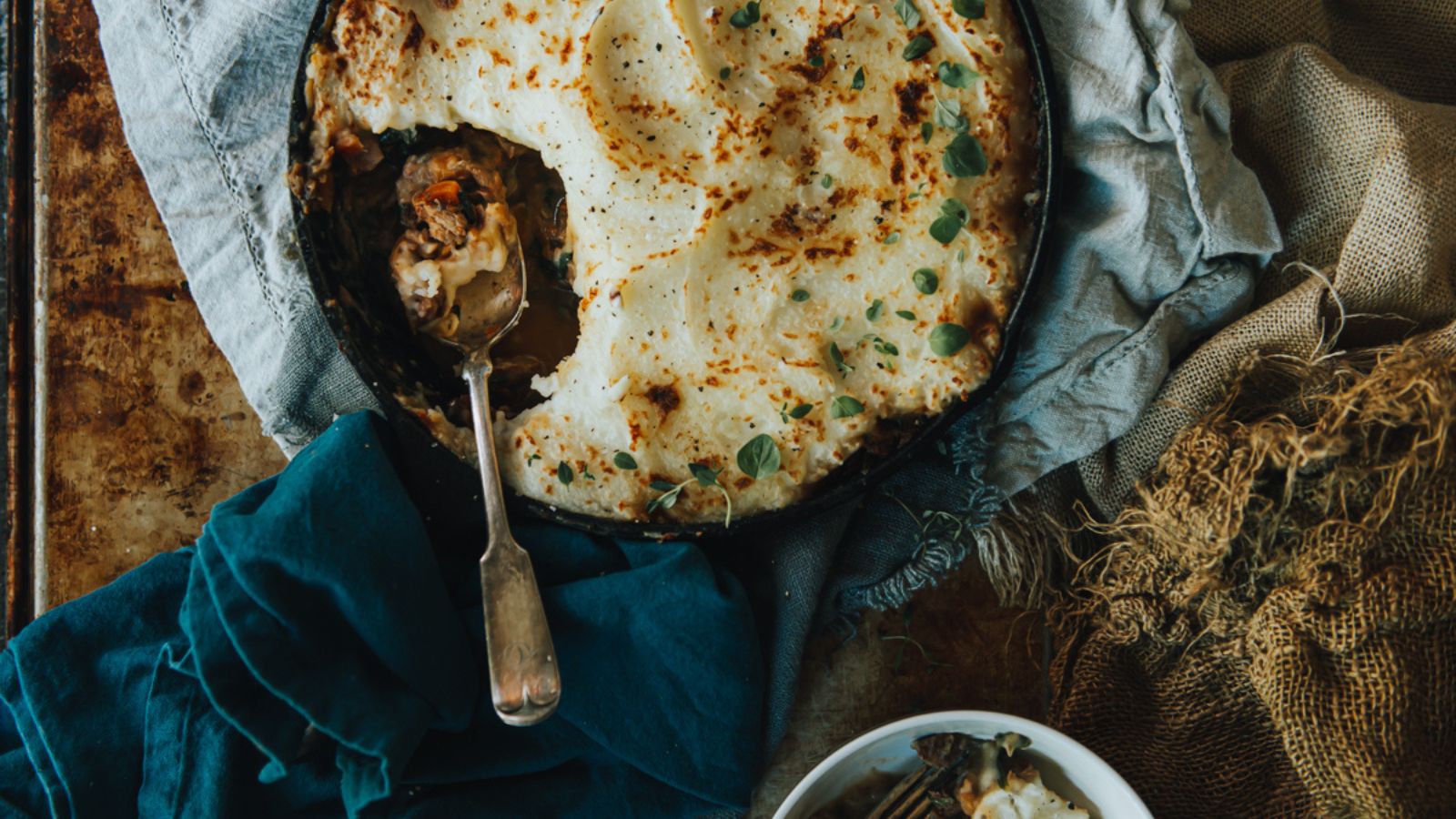 A pan of shepherd's pie with a serving spoon, surrounded by blue and burlap fabric. A small bowl with a portion of the pie is nearby.