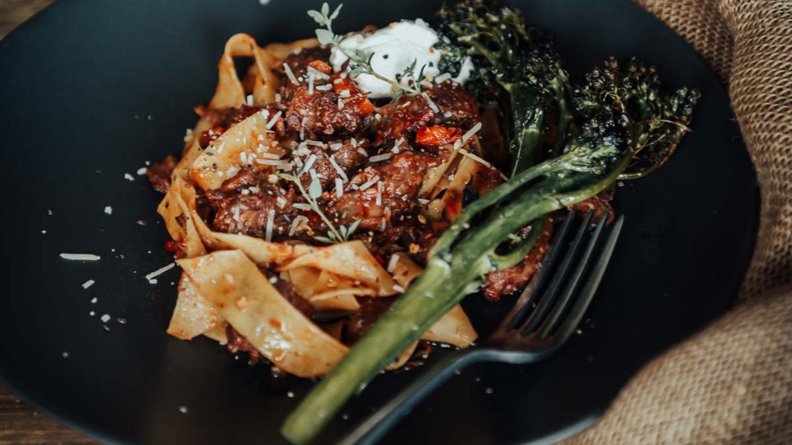 A black plate with pappardelle pasta, meat sauce, a dollop of cream, and two broccolini stalks, garnished with grated cheese, next to a fork.