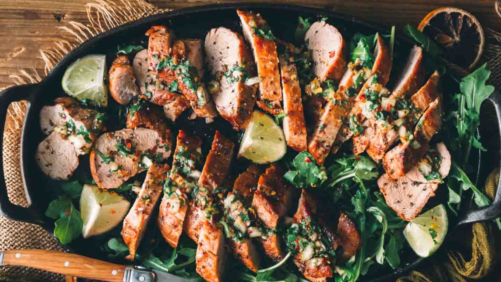 Sliced grilled meat garnished with herbs and lime, served on a bed of greens in a skillet.