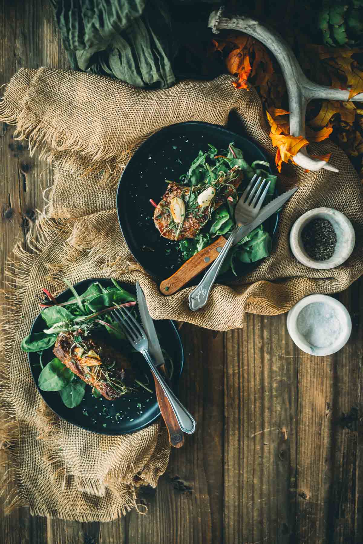 Two plates of boar chops served on a bed of greens with cutlery, placed on a rustic wooden table with burlap and autumn decorations.