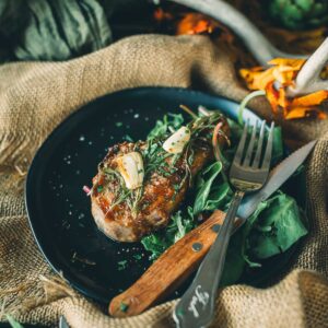 Seard wild boar chops garnished with garlic and herbs on a bed of greens, served on a black plate with a wooden-handled fork and knife.