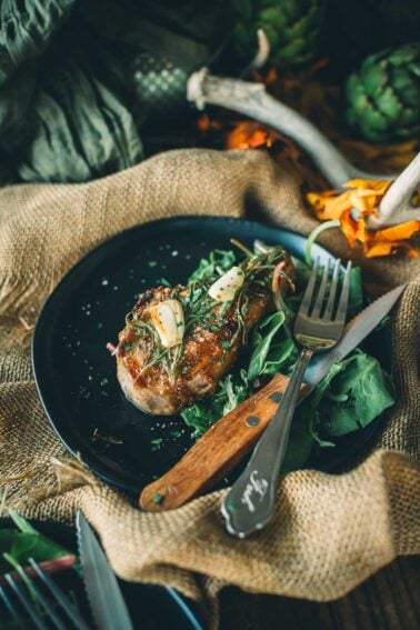 Seard wild boar chops garnished with garlic and herbs on a bed of greens, served on a black plate with a wooden-handled fork and knife.