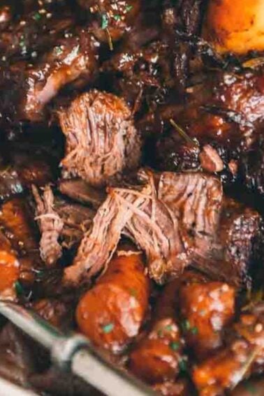 A close-up of a bowl of beef stew with carrots and a rich, dark sauce, garnished with herbs. A spoon is partially submerged in the dish.