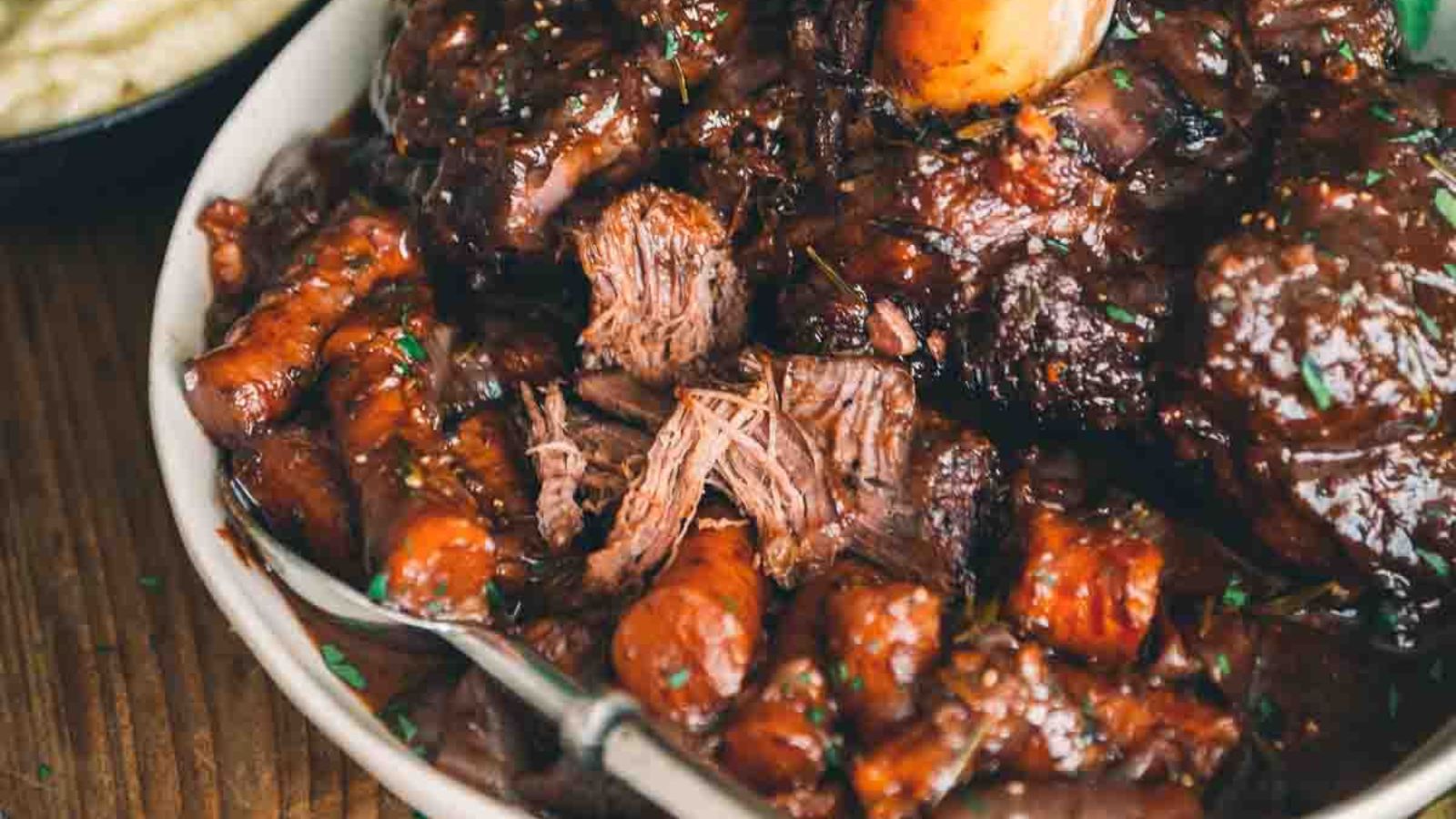 A close-up of a bowl of beef stew with carrots and a rich, dark sauce, garnished with herbs. A spoon is partially submerged in the dish.