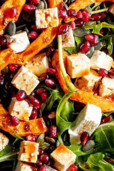 A salad with arugula, roasted pumpkin slices, feta cheese, pomegranate seeds, and pumpkin seeds.