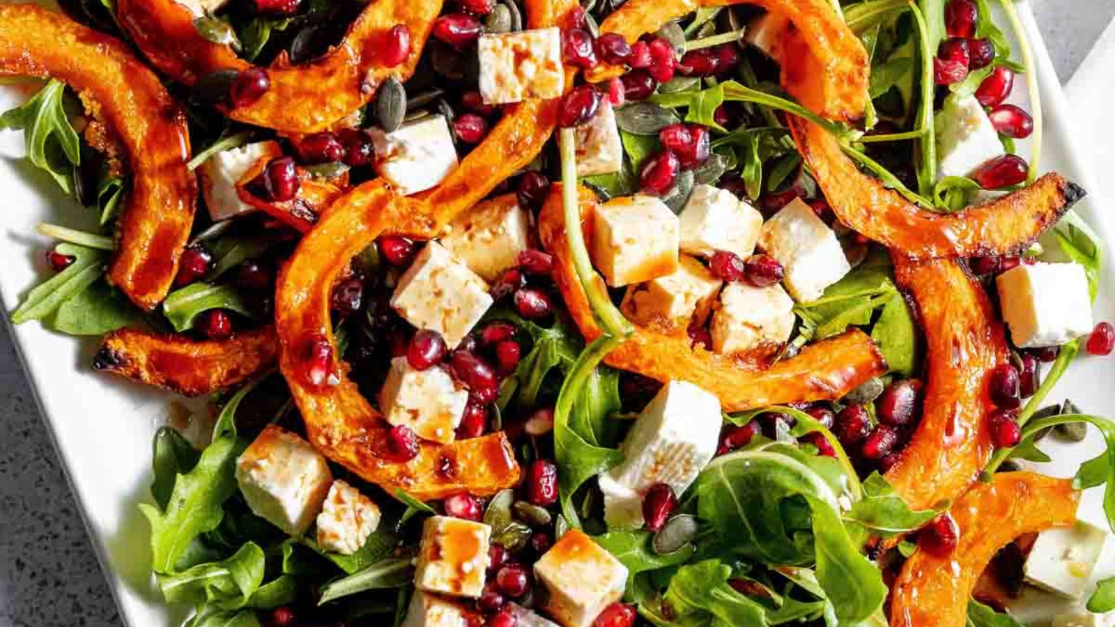 A salad with arugula, roasted pumpkin slices, feta cheese, pomegranate seeds, and pumpkin seeds.