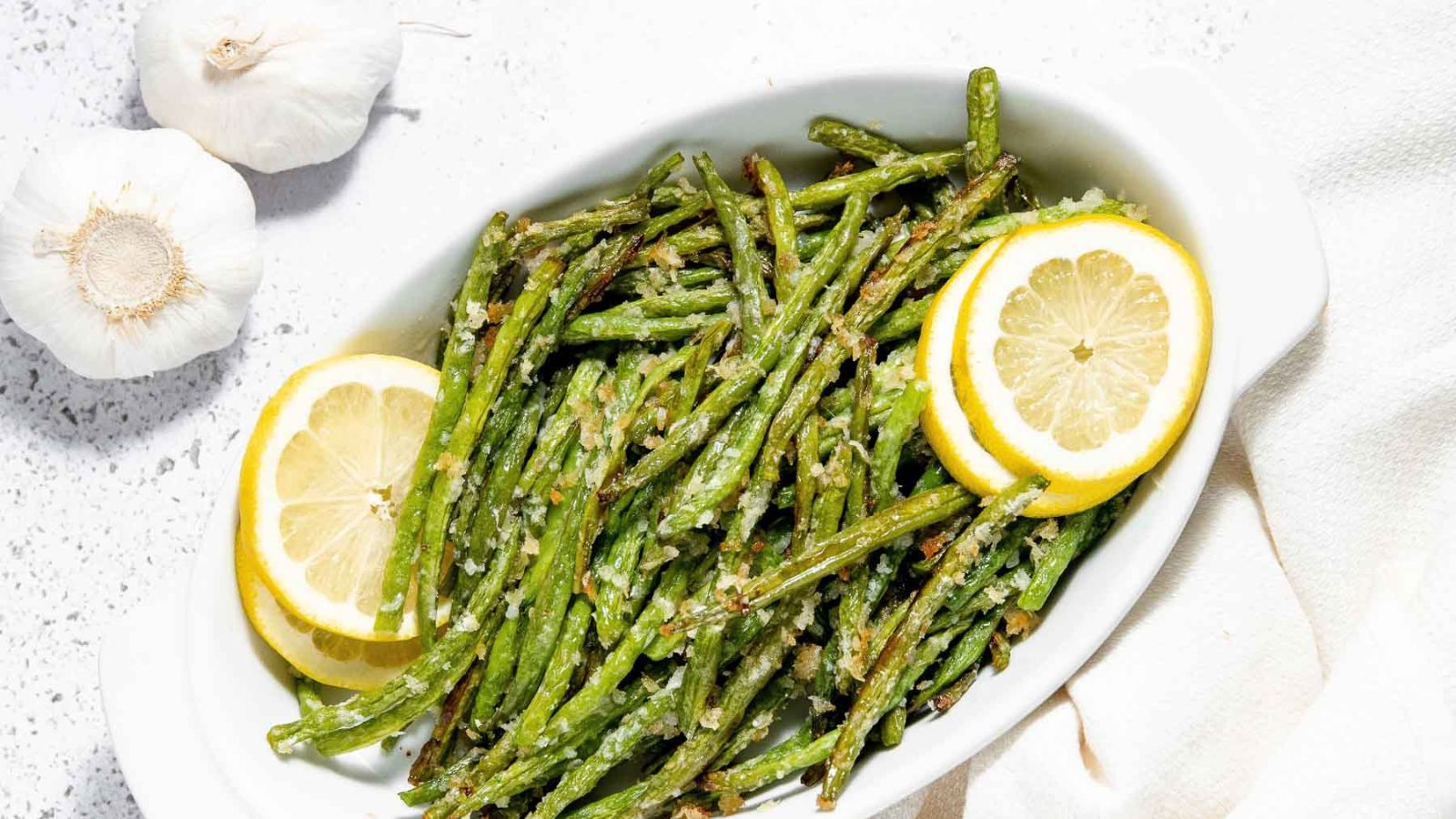 A white dish filled with seasoned green beans garnished with lemon slices. Two garlic bulbs are placed beside the dish.