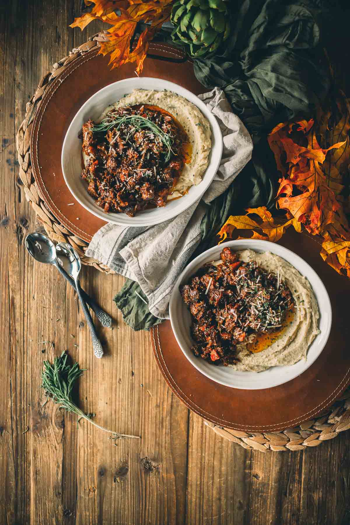 Two bowls of slow cooker beef shanks over roasted parsnip puree and herbs on a wooden table, surrounded by autumn leaves and artichokes, with a pair of spoons on the side.