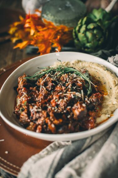 A bowl of rich shredded slow cooked beef shanks topped with shredded cheese and herbs, accompanied by creamy mashed potatoes, set on a rustic table with autumn decorations in the background.