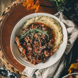 Bowl of slow cooked beef shanks with herbs on creamy polenta, garnished with rosemary, set on a woven mat with decorative leaves and artichoke.