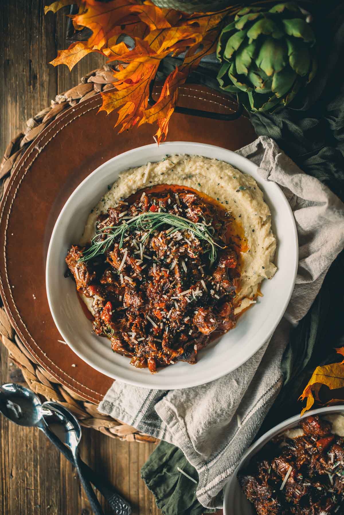 Bowl of slow cooked beef shanks with herbs on creamy polenta, garnished with rosemary, set on a woven mat with decorative leaves and artichoke.