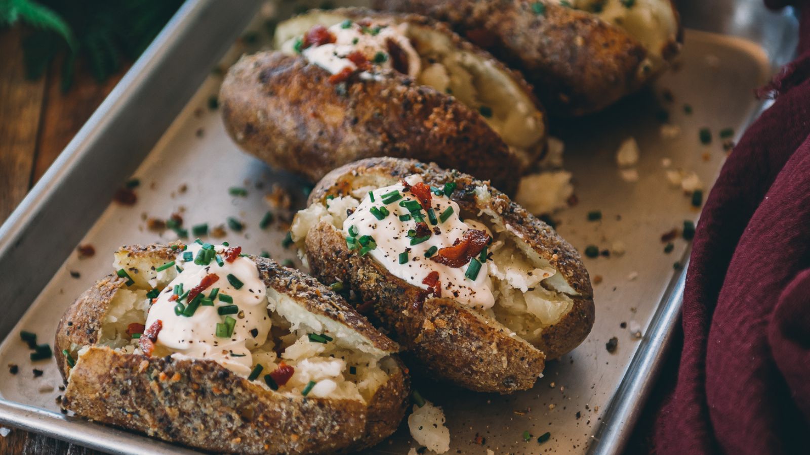 Baked potatoes topped with sour cream, chopped chives, and bacon bits on a baking tray.