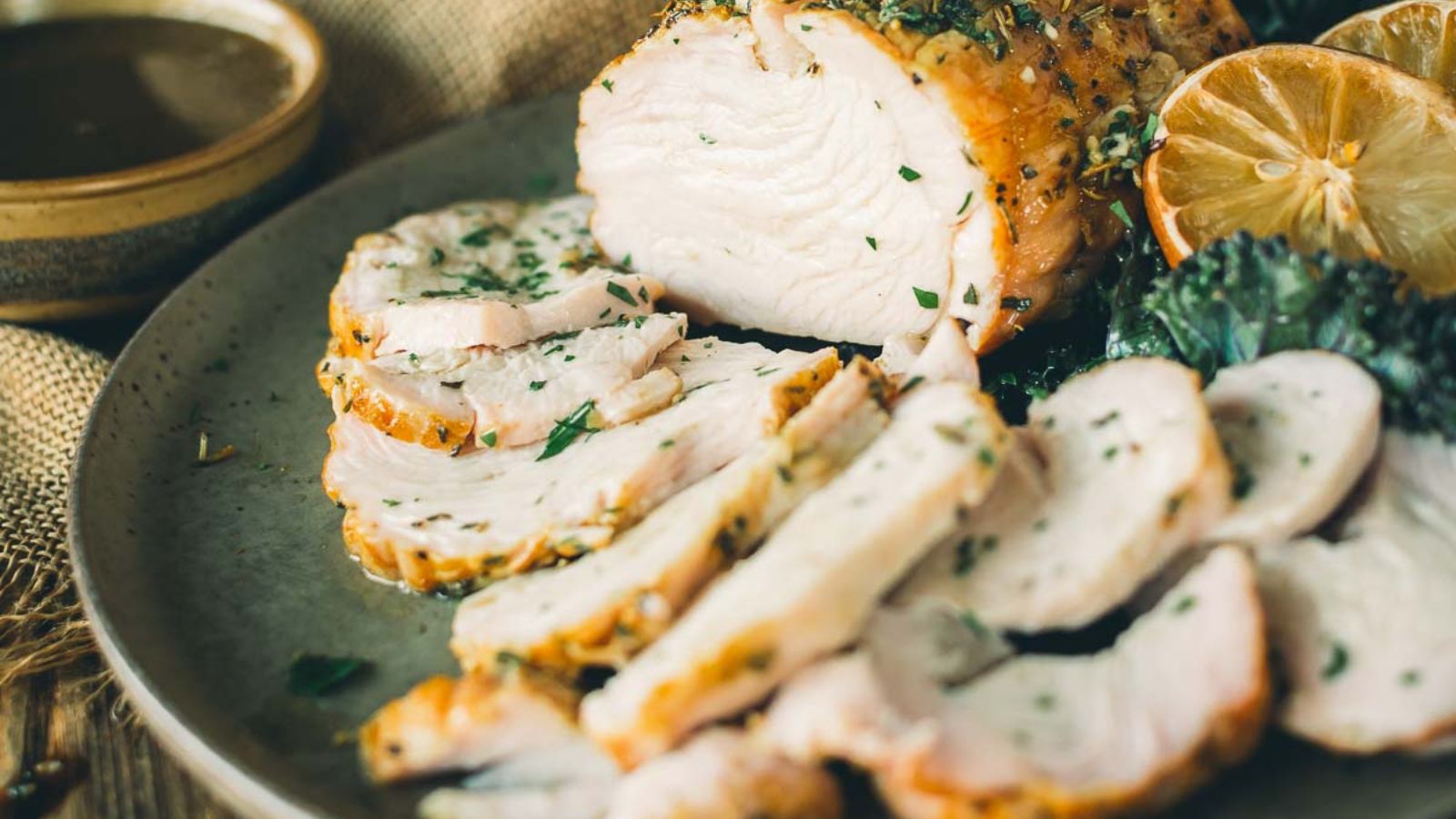 Sliced roasted turkey breast garnished with herbs on a plate, alongside lemon and greens.