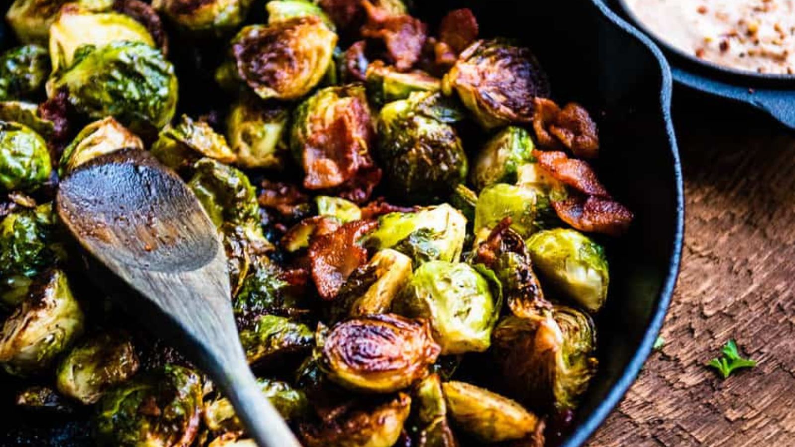 A cast iron skillet with roasted Brussels sprouts and crispy bacon, accompanied by a wooden spoon.