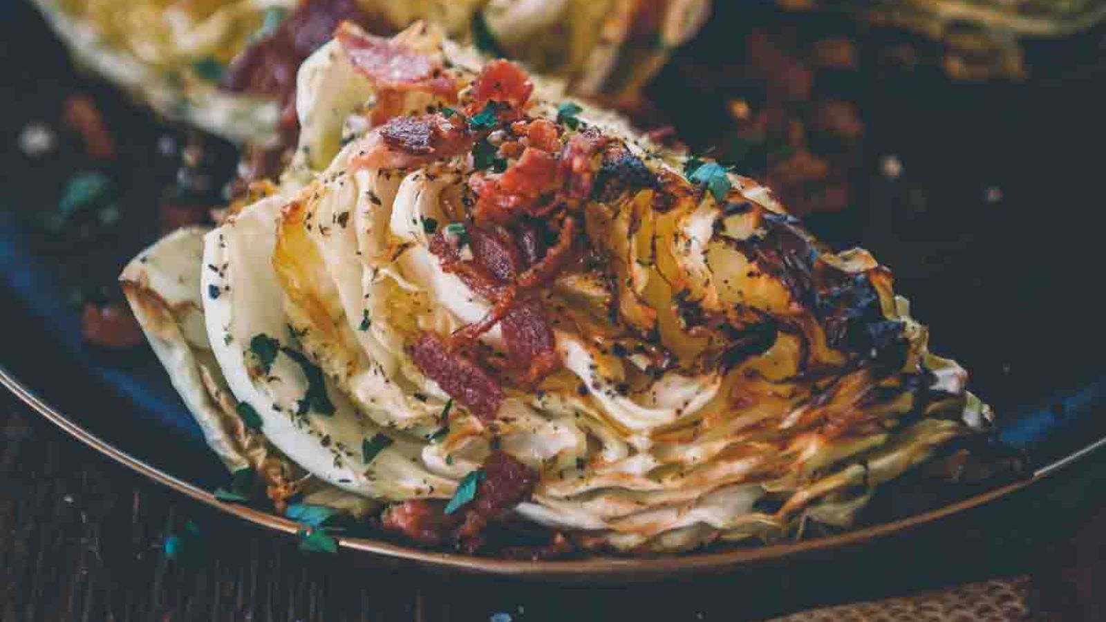 Grilled cabbage wedge topped with crispy bacon pieces and herbs on a dark plate.