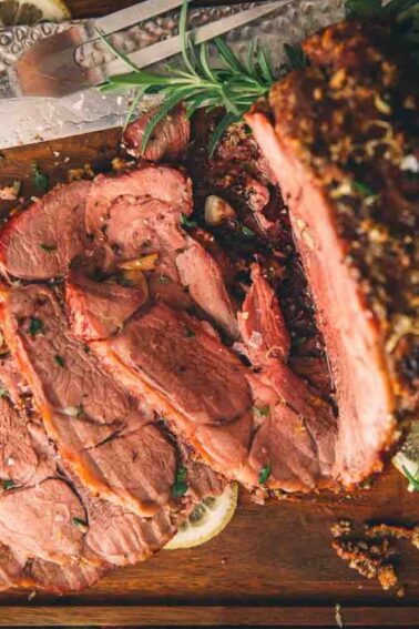Sliced roast beef on a wooden cutting board with a knife, accompanied by herbs and lemon slices.