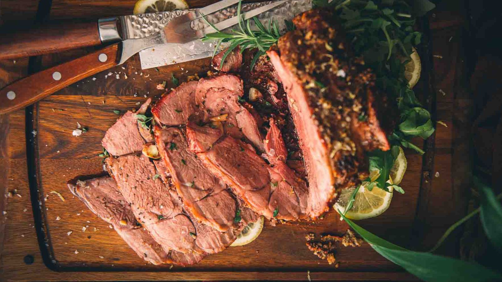 Sliced roast beef on a wooden cutting board with a knife, accompanied by herbs and lemon slices.