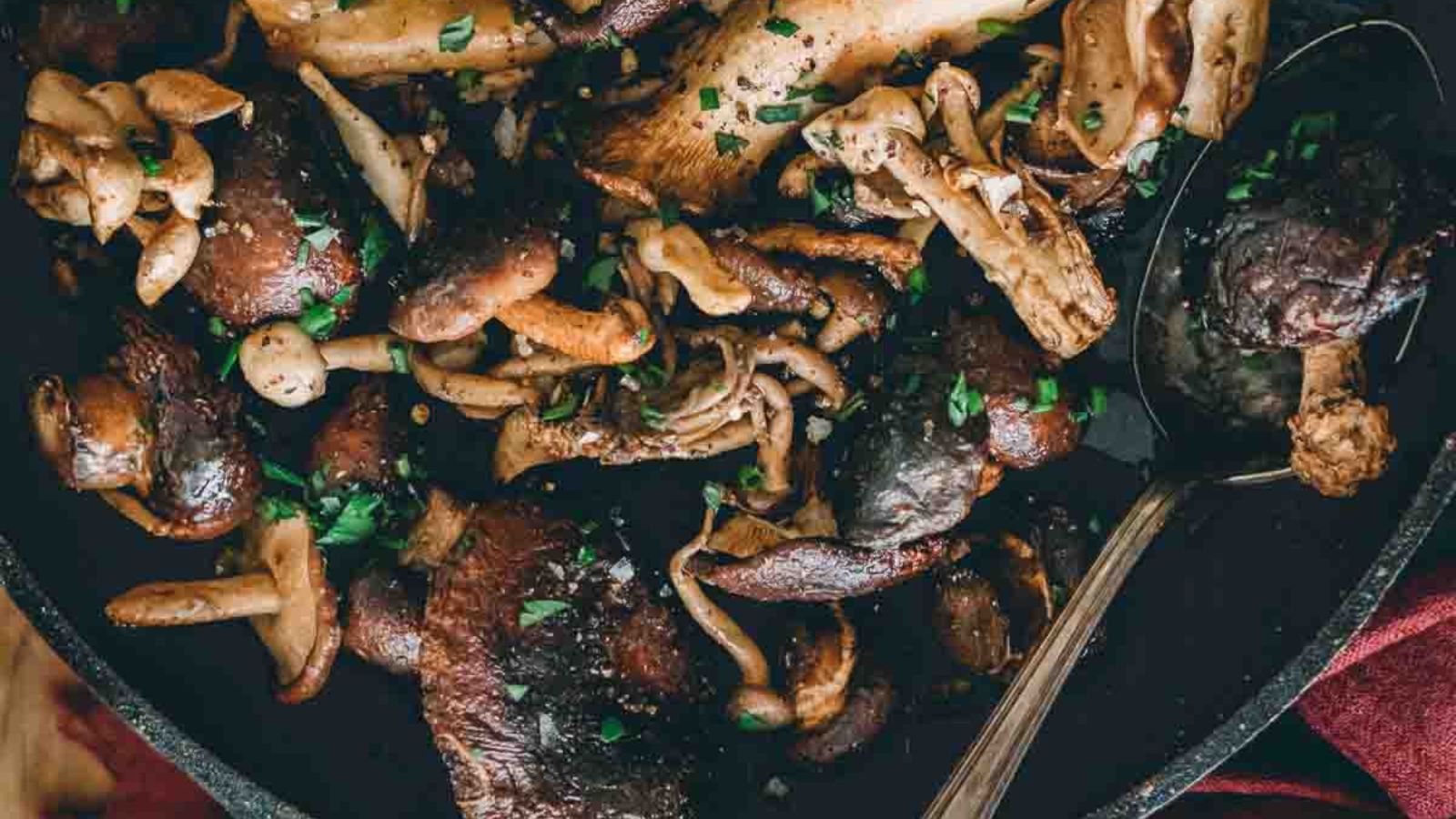 A variety of mushrooms are cooked in a pan, accompanied by chopped herbs and a large serving spoon.