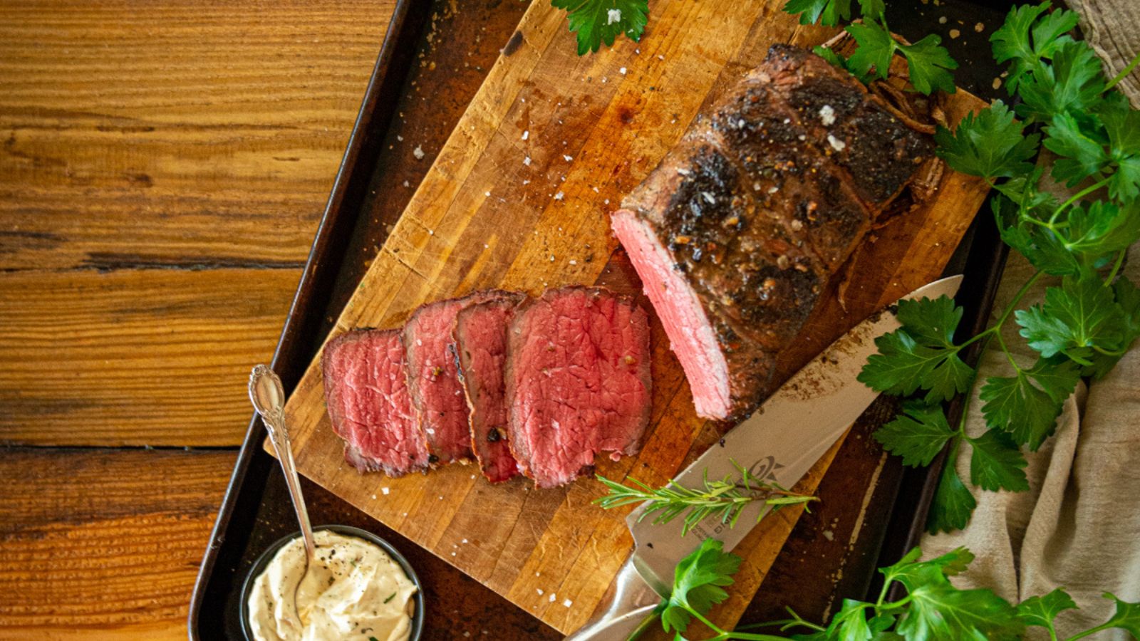 Sliced roast beef on a wooden cutting board with parsley and a knife, served with a small bowl of creamy sauce on the side.