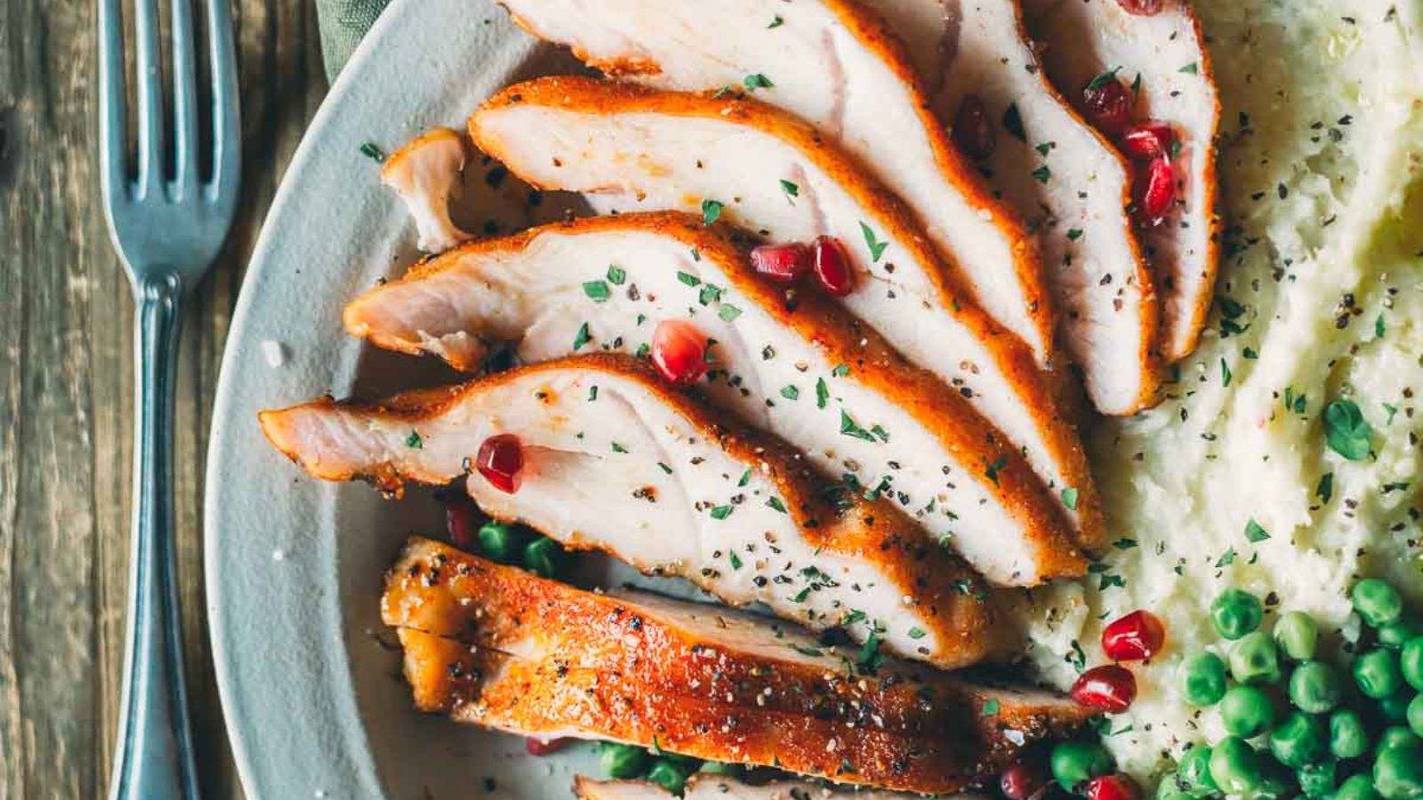 Sliced smoked turkey tenderloin with pomegranate seeds and herbs, served alongside mashed potatoes and peas on a plate.