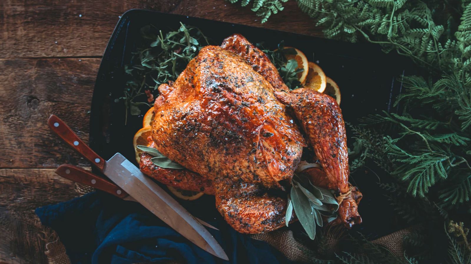 Roasted chicken garnished with herbs and orange slices on a dark platter, with two knives placed nearby.