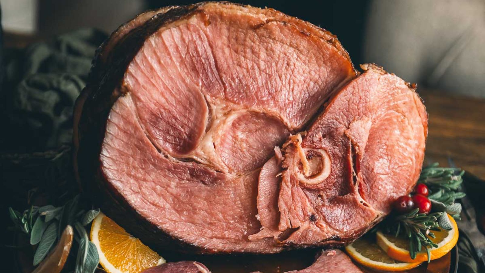 Close-up of a sliced ham garnished with orange slices, herbs, and cranberries on a wooden surface.