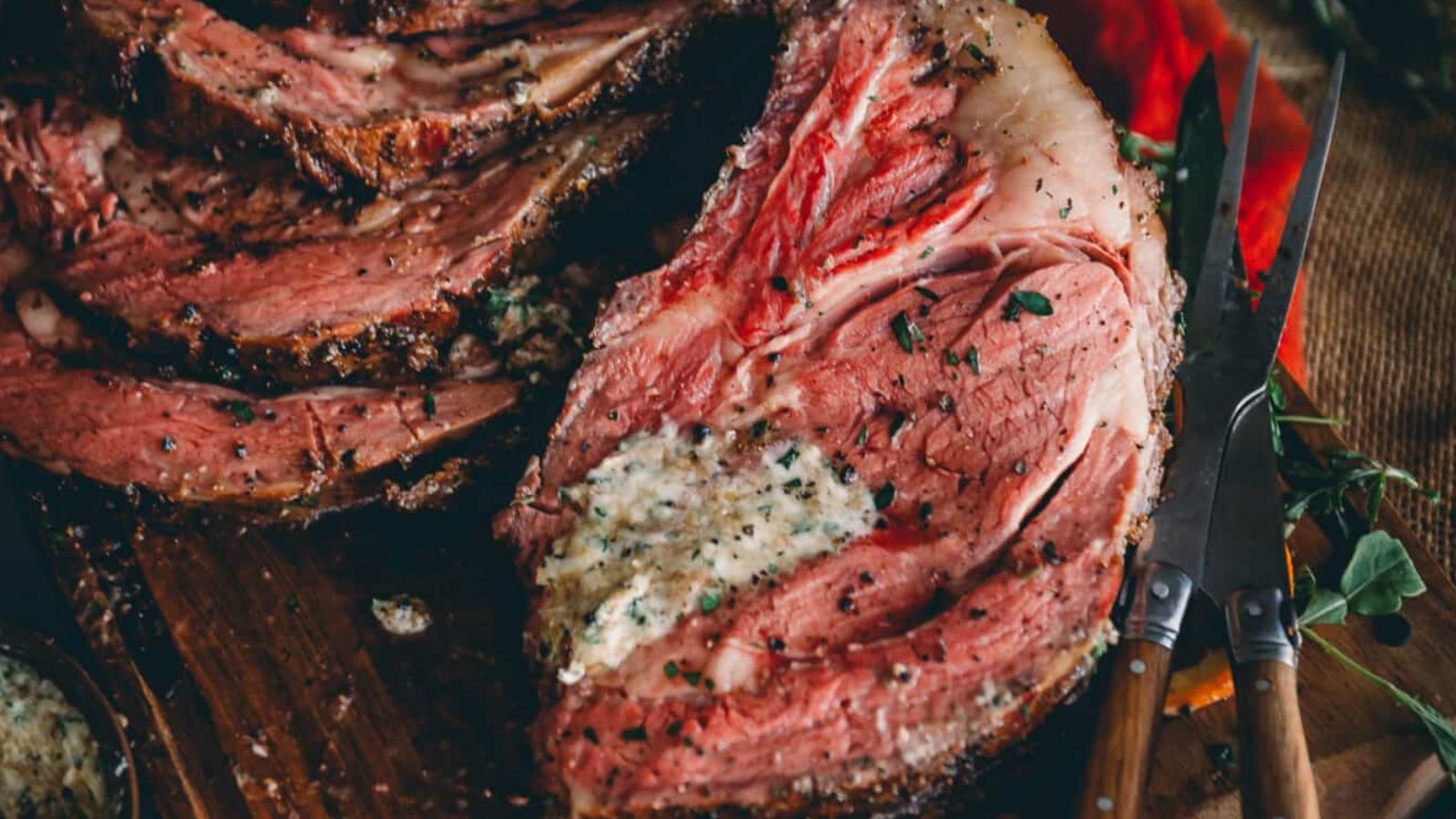 Close-up of a sliced, seasoned prime rib roast on a wooden board, accompanied by a knife and fork.