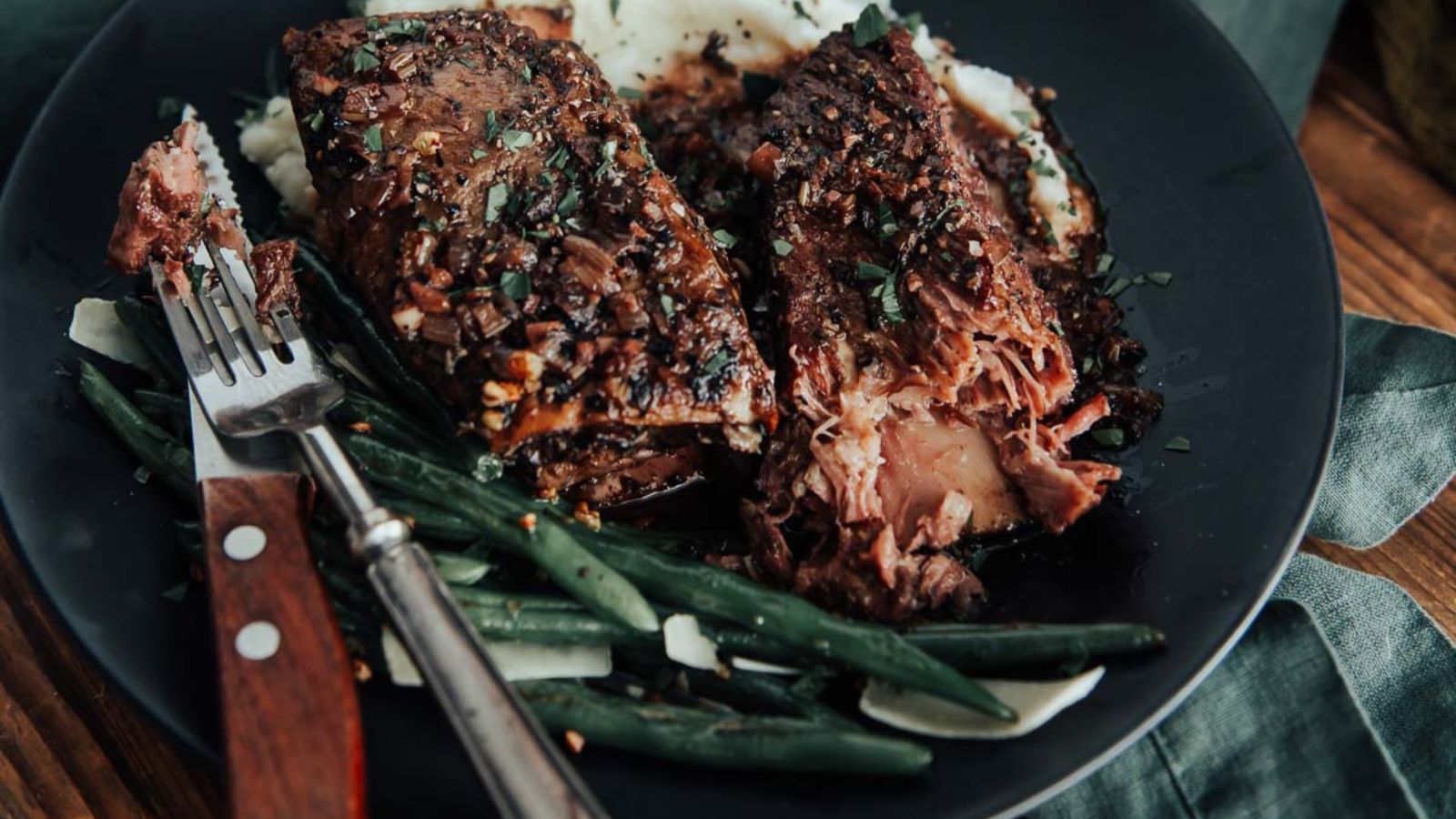 A plate with sliced short ribs, mashed potatoes, and green beans. A fork rests on the side.