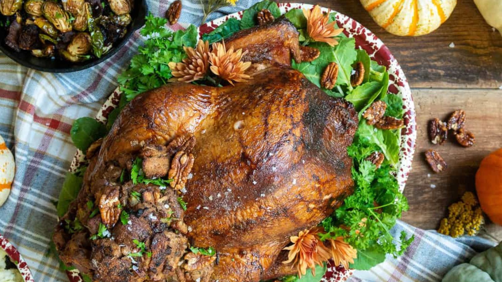 Roasted turkey with stuffing on a platter, garnished with herbs and nuts. Surrounded by a checkered cloth and autumn decorations.