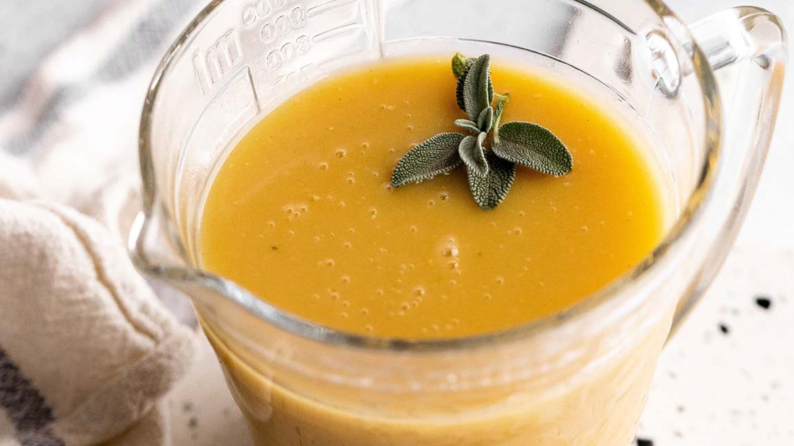 A glass measuring cup filled with smooth yellow gravy, topped with two green sage leaves. A cloth is partially visible nearby.