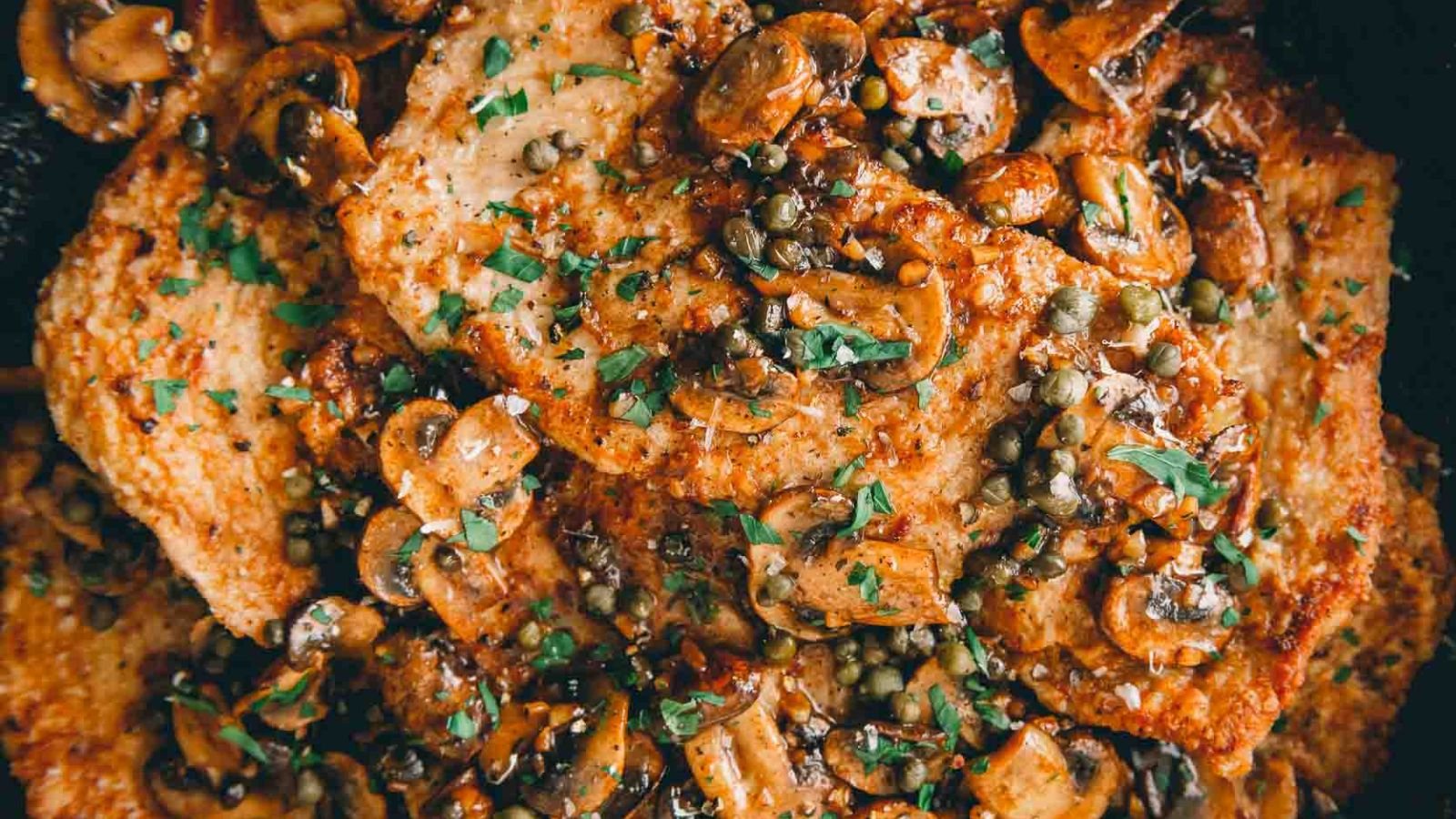 Close-up of chicken marsala topped with sautéed mushrooms, capers, and herbs.